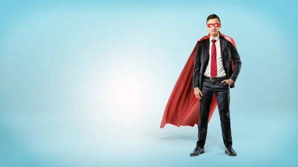 Un uomo d'affari sicuro di sé con un mantello rosso e una maschera in piedi con una mano in tasca su sfondo blu . — Foto Stock
