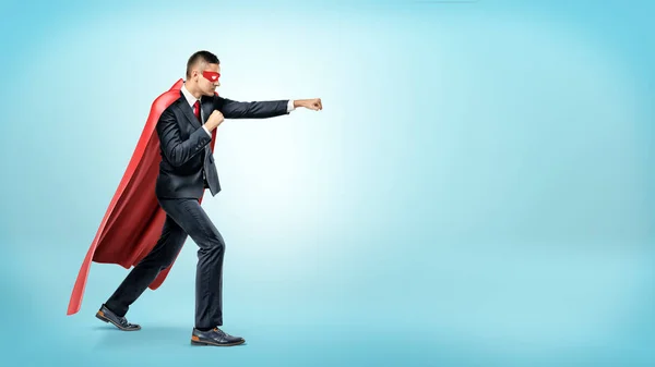 Un uomo d'affari con un mantello rosso fluente da supereroe e una maschera che lancia pugni contro un nemico invisibile su sfondo blu . — Foto Stock