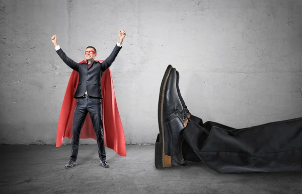 En leende affärsman i en superhjälte röd cape med händerna upp i framgång motion stående bredvid en gigantisk mänsklig fot. — Stockfoto