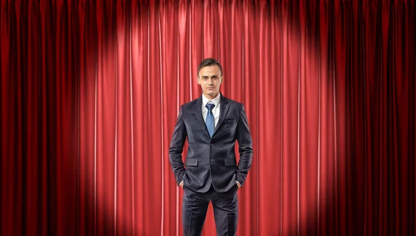 A confident businessman standing in front view in the spotlight with a red stage curtain behind him.