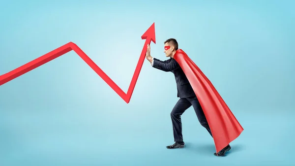Podnikatel v červené tekoucí cape snaží ohnout červený Statistika šipku nahoru na modrém pozadí. — Stock fotografie