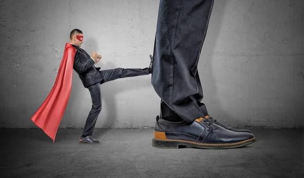 A tiny businessman in a red cape and an eyemask kicking at a retreating giant foot. — Stock Photo, Image