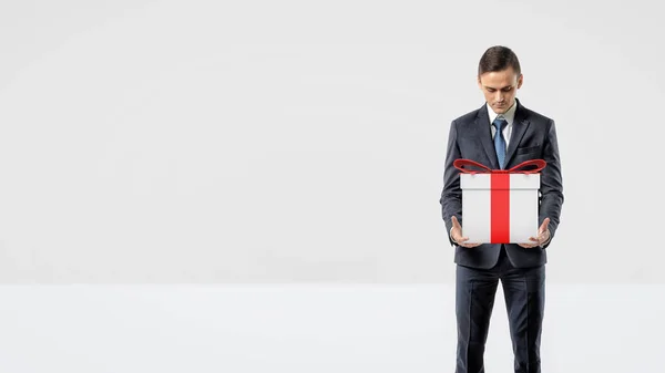 A businessman holding a gift box with a red bow in his arms looking down at it. — Stock Photo, Image