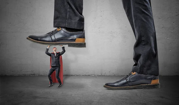 Un pequeño hombre de negocios con capa roja de superhéroe tratando de sostener una bota gigante pisándole . — Foto de Stock