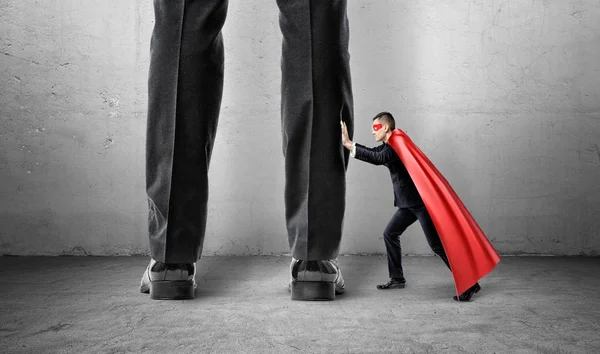 Un pequeño hombre de negocios en una capa de superman tratando de alejar las piernas de un hombre gigante . — Foto de Stock