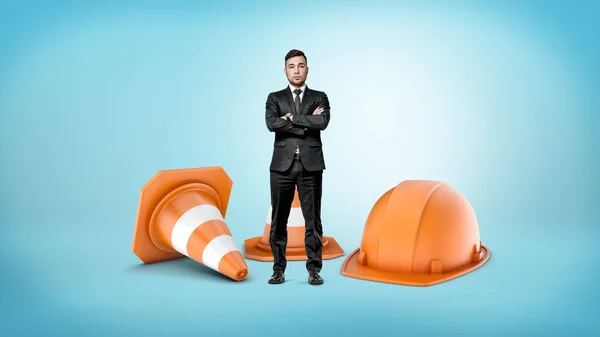 Un pequeño hombre de negocios con las manos cruzadas de pie junto a gigantes conos de tráfico a rayas y un casco naranja . — Foto de Stock