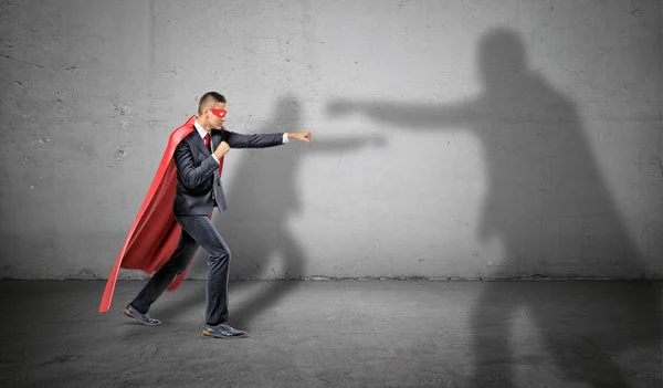 Un hombre de negocios con una capa roja y una máscara luchando contra una sombra de otro hombre más grande sobre un fondo de hormigón . — Foto de Stock