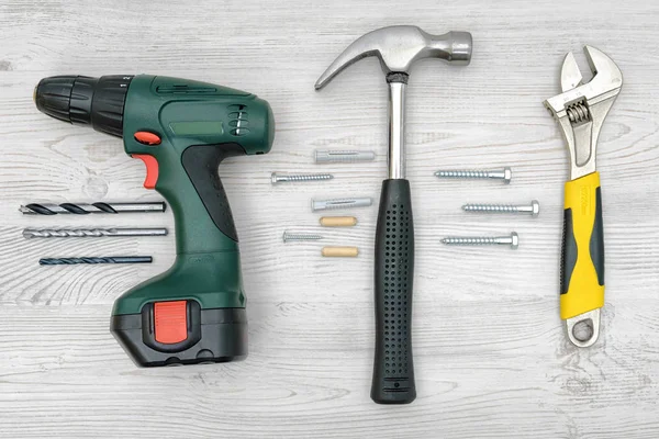 A drill, a hammer and a wrench on wooden background with nails, dowels, bits and screw bolts between them. — Stock Photo, Image