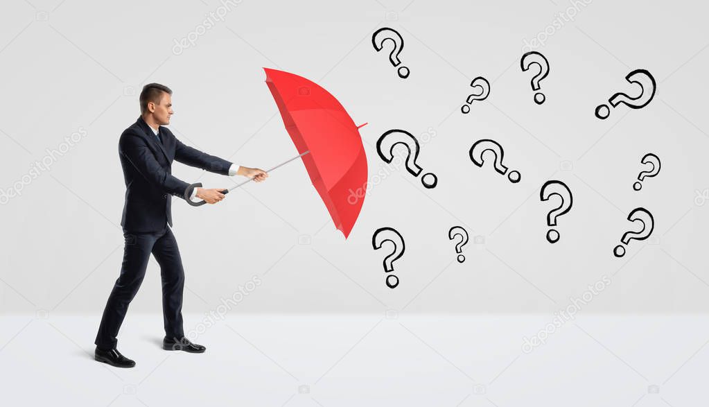 A businessman covering himself with an open red umbrella from many black drawn question marks.