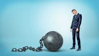 A small sad businessman looking down to a giant iron ball and chain on blue background. clipart