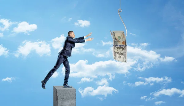 Een kleine zakenman staande op een betonnen kolom en vangen van een dollarbiljet gevangen op een metalen haakje. — Stockfoto