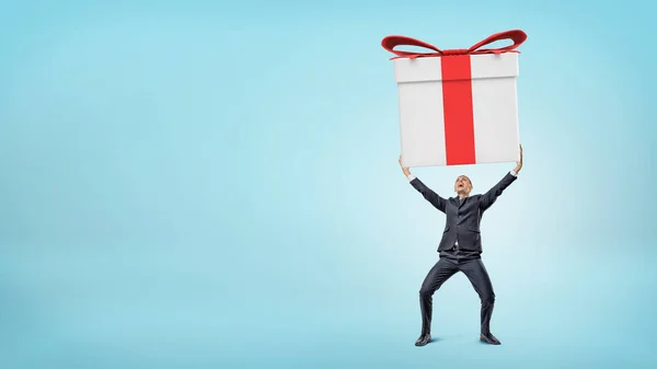 Un pequeño hombre de negocios feliz de pie sobre fondo azul y sosteniendo una caja de regalo gigante con ambas manos . — Foto de Stock