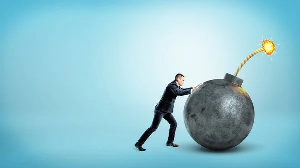 Un pequeño hombre de negocios a la vista empujando una bomba redonda gigante con un fusible encendido sobre fondo azul . —  Fotos de Stock