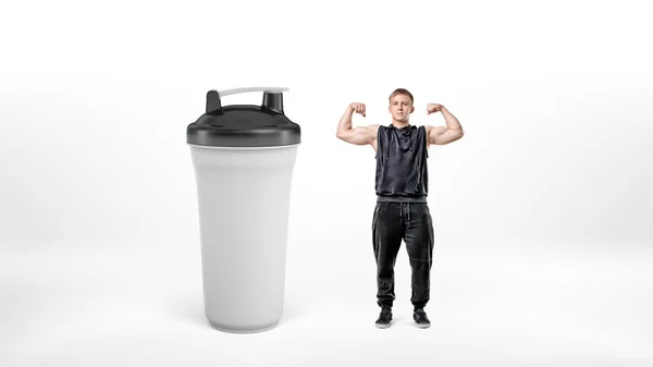 Ein kleiner muskulöser Mann mit weißem Bizeps, der neben einem riesigen weißen Sport-Shaker steht. — Stockfoto