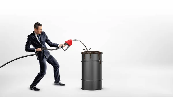 A businessman holds a gas nozzle and overfills a black barrel with oil. — Stock Photo, Image