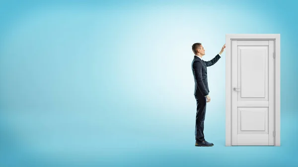 Un hombre de negocios sobre fondo azul toca un marco de puerta blanco con una puerta cerrada dentro . — Foto de Stock