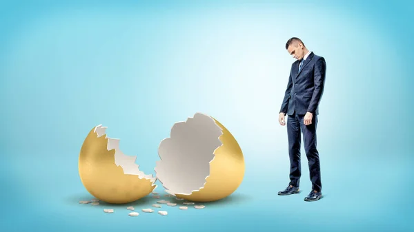 A sad businessman on blue background looks down on a giant broken golden egg.