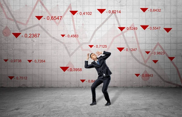 Испуганный бизнесмен согнулся у бетонной стены с красными биржевыми индексами и падающими статистическими линиями . — стоковое фото