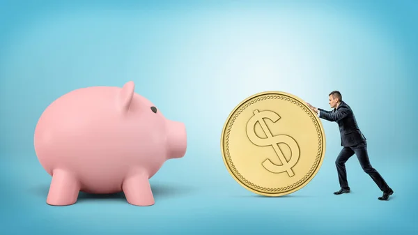 Een kleine zakenman rolt een enorme gouden dollar munt in de richting van een gigantische piggy bank. — Stockfoto