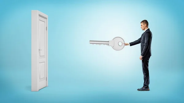 Бизнесмен держит гигантский серебряный ключ, готовый открыть белую изолированную дверь на синем фоне. . — стоковое фото