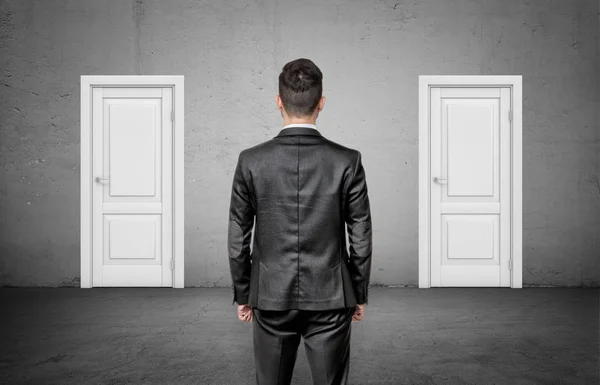 Un hombre de negocios con la espalda vuelta se encuentra entre dos puertas blancas cerradas idénticas . — Foto de Stock