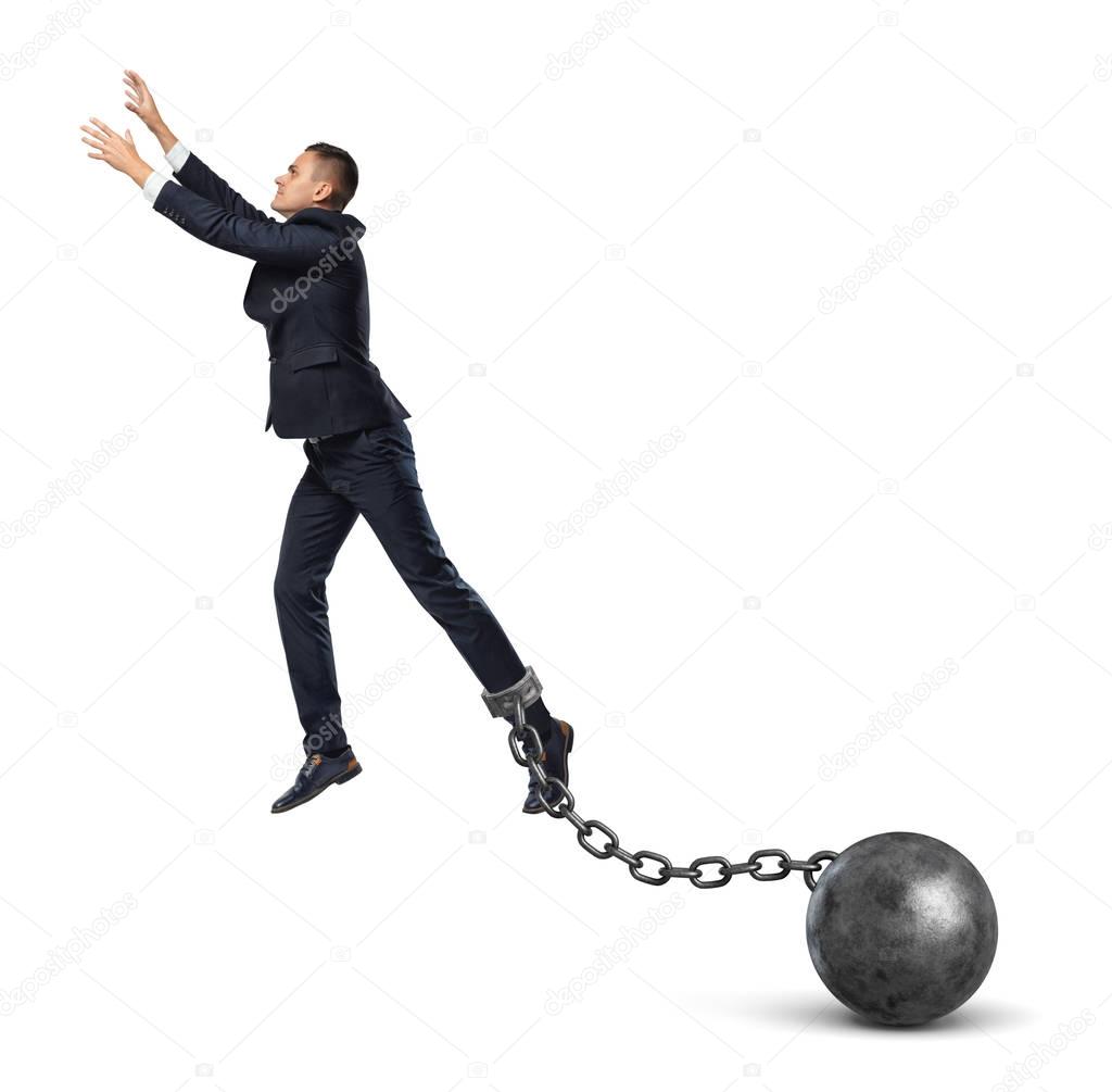 A businessman shackled to a huge iron ball leaps up and tries to catch something above.