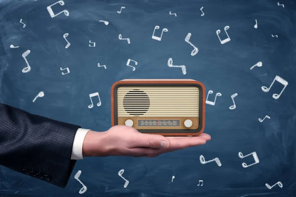 A small retro radio set with brown housing and yellow front stands on a businessmans palm on blackboard background.