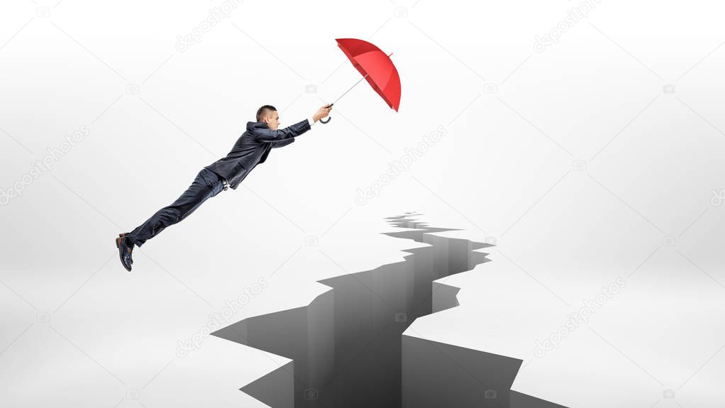 A businessman uses a large red umbrella to fly over a long earthquake crack on white background.