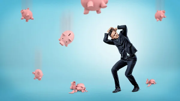 Ein Geschäftsmann versteckt seinen Kopf vor vielen Sparschweinen, die auf den Boden fallen und knacken. — Stockfoto
