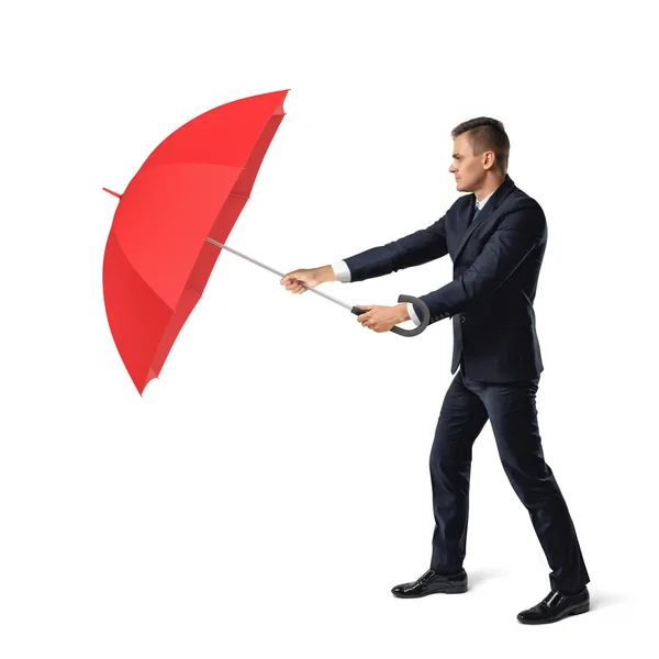 Ένας νεαρός επιχειρηματίας βρίσκεται σε λευκό φόντο και κρύβει από σκληρό περιβάλλον πίσω από μια ανοιχτή κόκκινη ομπρέλα. — Φωτογραφία Αρχείου