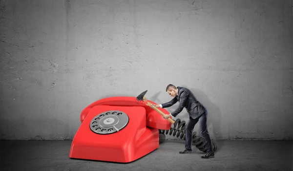 Um pequeno empresário não consegue destruir um grande telefone retro vermelho com um martelo quebrado . — Fotografia de Stock