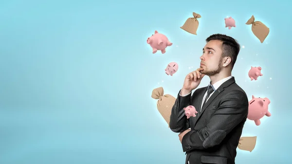 Een jonge zakenman diep in gedachte met kleine piggy banken en geld zakken vliegen rond zijn hoofd baard. — Stockfoto