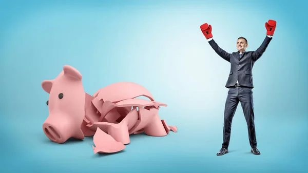 Een gelukkig zakenman heeft zijn handen opgewekt in overwinning terwijl hij naast een gebroken piggy bank staat. — Stockfoto