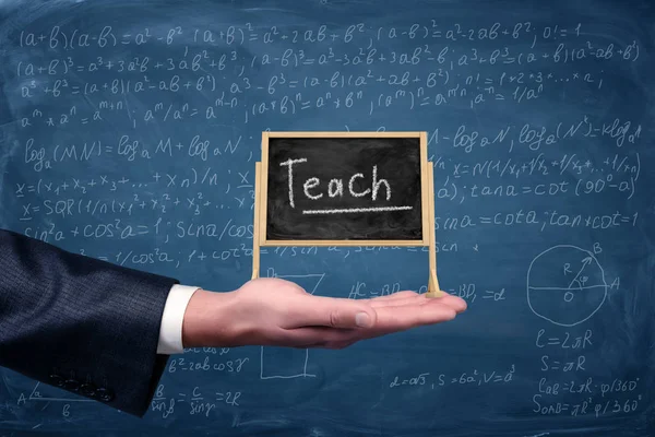 De hand van een businessmans houdt een kleine ezel schoolbord met een woord Teach daarop met vergelijkingen op de achtergrond. — Stockfoto