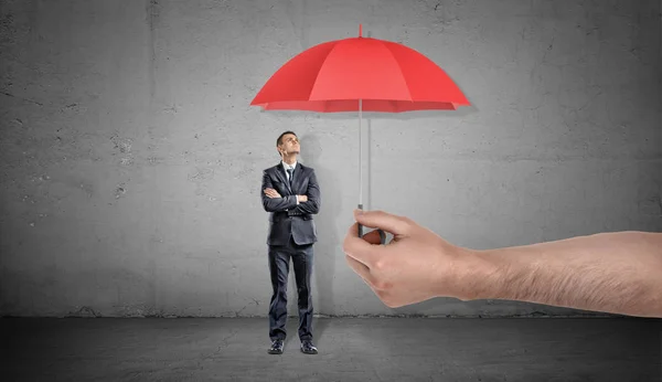 Een kleine zakenman staat op een rode open paraplu aangeboden door een gigantische mannenhand opzoeken. — Stockfoto