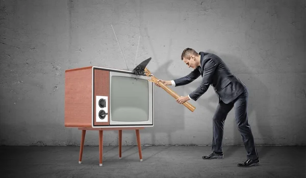 Um homem de negócios em fundo de concreto não consegue esmagar um velho aparelho de TV retro com um martelo quebrado . — Fotografia de Stock