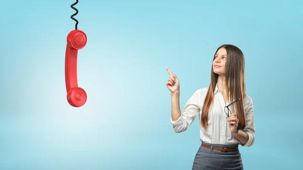 A beautiful businesswoman points to a large red phone receiver hanging from a cord.