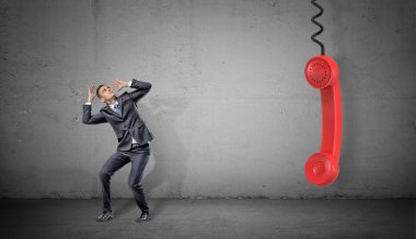 Aşağı asılı bir büyük kırmızı retro telefon işleyicisi yakınındaki beton zemin üzerine küçük bir korkmuş işadamı.