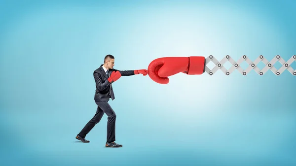 Een kleine ondernemer in sport handschoenen stoten een gigantische rode Bokshandschoen op de arm van een metalen schaar. — Stockfoto