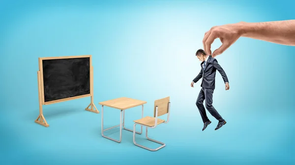 Um empresário minúsculo pendurado em uma mão gigante sobre fundo azul com mobiliário escolar e um quadro-negro . — Fotografia de Stock
