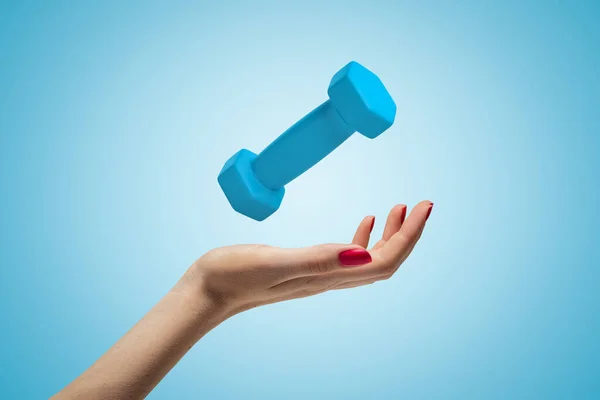 Side closeup of womans hand facing up and levitating small bright-blue dumbbell on light-blue gradient background.
