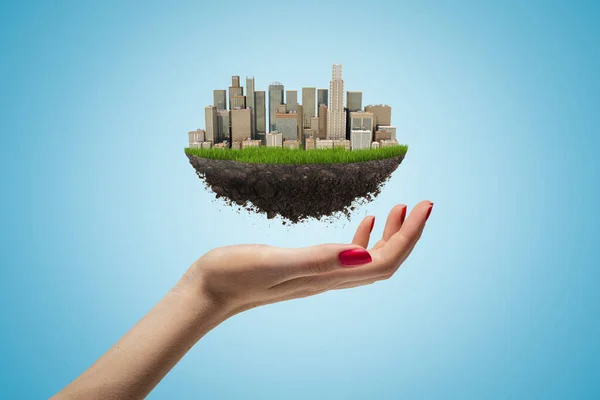 Side closeup of womans hand facing up and levitating small piece of land with modern city on it on light blue background. — Stock Photo, Image