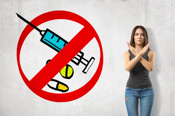 Serious young girl with hands crossed, standing against wall with picture of syringe and some meds enclosed in red prohibition sign. — Stock Photo, Image