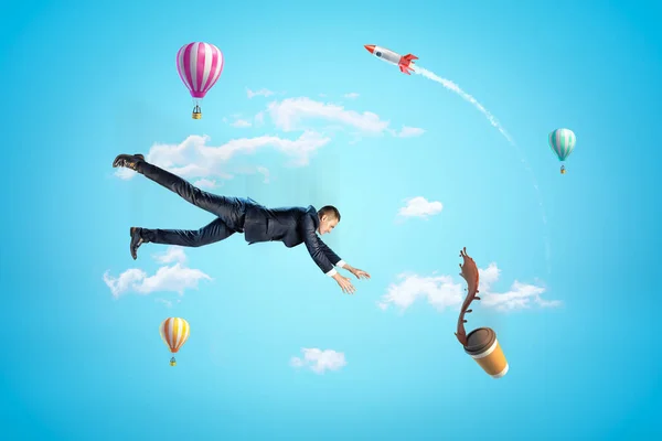 Jovem empresário no céu caindo tentando pegar xícara de café de papel que está derramando café no ar com balões de ar quente e foguete no fundo . — Fotografia de Stock