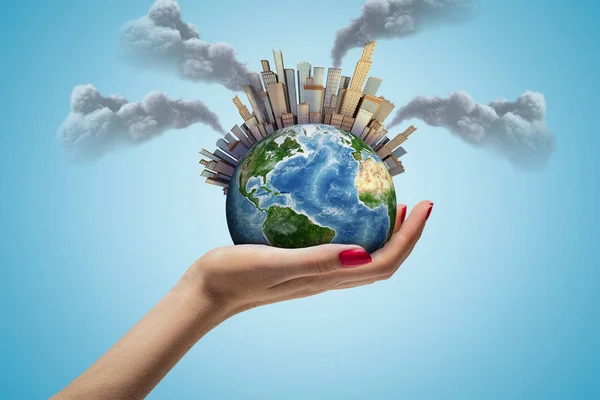 Side closeup of womans hand facing up and holding small Earth with skyscraper city on it emitting plumes of grey smoke in air on light-blue background. — Stock Photo, Image