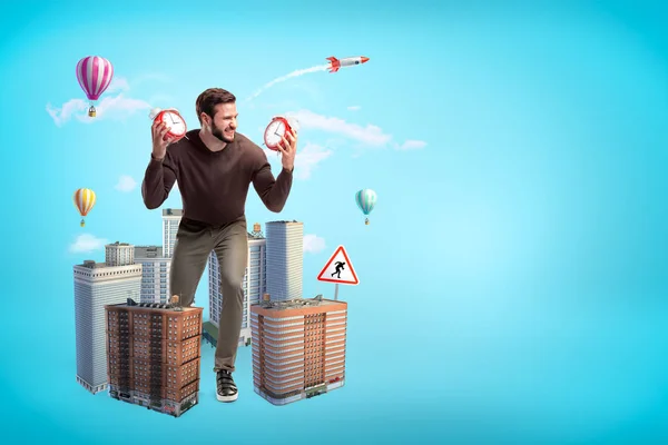 Angry young man in casual clothes holding two alarm clocks with city skyscrapers and hot air balloons on blue background — 스톡 사진