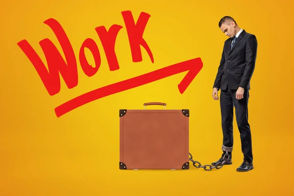 Droevige, vermoeide zakenman in halve bocht geketend aan bruine koffer op amberkleurige achtergrond met grote rode titel Werk. — Stockfoto