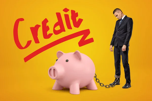 Droevige zakenman geketend aan roze spaarvarken bank met Credit teken hierboven op gele achtergrond. — Stockfoto