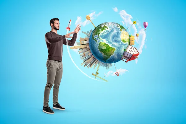 Young man standing on light blue background trying to touch small Earth which is floating in air, with big city, house and road signs on it. — Stock Photo, Image