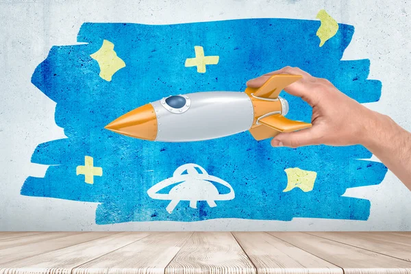 Mans hand spelen met zilver en oranje ruimte raket tegen muur met vaag schilderij van blauwe sterrenhemel en kleine UFO erin. — Stockfoto